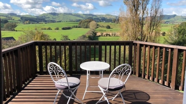 A wooden deck with two white metal chairs and a small round table, overlooking a green valley with hills and trees.