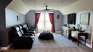 A spacious room with a ceiling fan, a large window with red curtains, a seating area with leather chairs, a small ottoman, and a TV on a wooden stand.