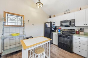 A compact kitchen with white cabinets, a black refrigerator, a microwave, and an oven. There is a small table with a stool and a metal shelf with a yellow bowl.