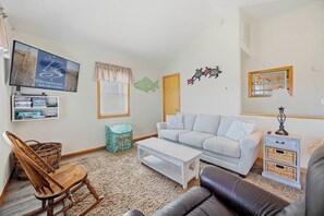A living room with a sofa, a coffee table, a wooden rocking chair, a television mounted on the wall, and a small side table with a lamp. - Six Pack | 1169 ft from the beach | Dog Friendly, Private Pool, Hot Tub | Nags Head (Nags Head)
