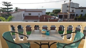 A balcony with two turquoise chairs and a table, overlooking a coastal area with buildings and a clear sky.
