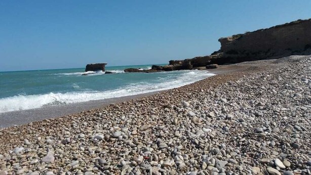 A pebbled beach with waves, a rocky cliff, and clear skies.
