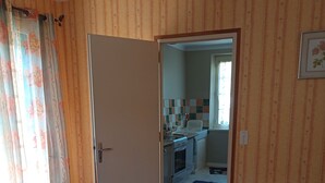 Une pièce avec un plancher en bois, un tapis à motifs, un lit, une porte donnant sur une cuisine avec un mur recouvert de carreaux, et un radiateur.