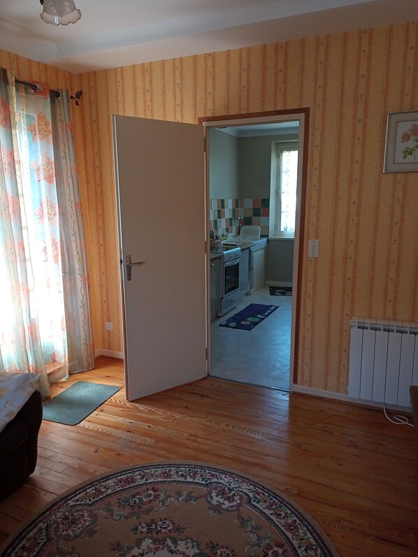 A room with wooden flooring, a patterned rug, a bed, a door leading to a kitchen with a tiled backsplash, and a radiator.
