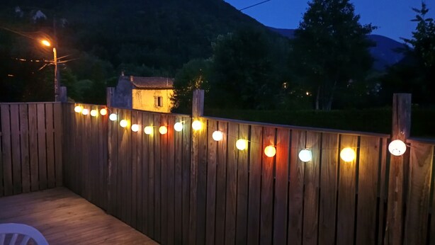Une terrasse en bois illuminée de guirlandes lumineuses, une chaise en plastique blanche et une maison en arrière-plan.