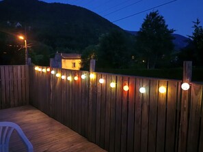 A wooden deck with string lights, a white plastic chair, and a house in the background.