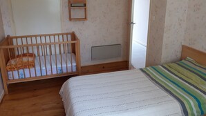 Une chambre avec un berceau en bois, un lit recouvert d’une couverture à rayures, un radiateur et une porte donnant sur une autre pièce.