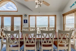 Uma sala de jantar com vista para o oceano, cadeiras de madeira, um ventilador de teto e um aviso com as regras da casa de praia.