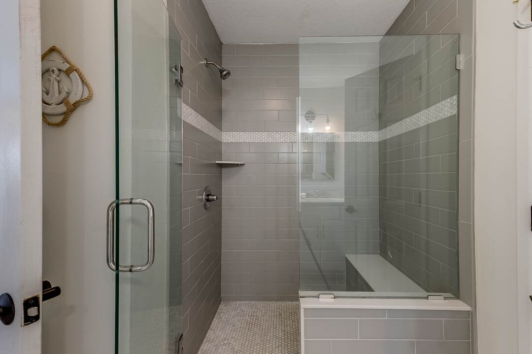 Une douche fermée par des parois en verre, avec des carreaux gris et une porte blanche dotée d’une poignée.