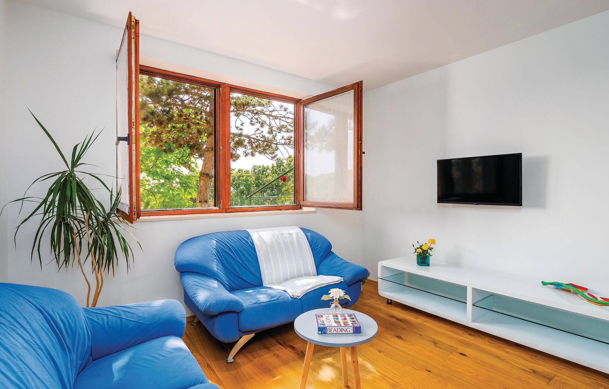 A modern living room with a blue sofa, a small round table with a book, a TV mounted on the wall, and a large window with a view of trees.