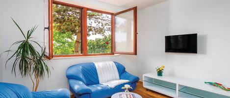 A modern living room with a blue sofa, a small round table with a book, a TV mounted on the wall, and a large window with a view of trees.
