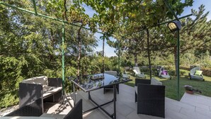 Een terras buiten met rieten meubilair, een tafel met glazen blad en een pergola begroeid met klimplanten.