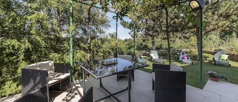 En udendørs patio med fletmøbler, et glasbord og en pergola dækket med slyngplanter.