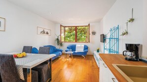 Een moderne woonkamer met een eethoek, een groot raam met uitzicht op bomen, een blauwe bank en een keuken met een koffieapparaat.