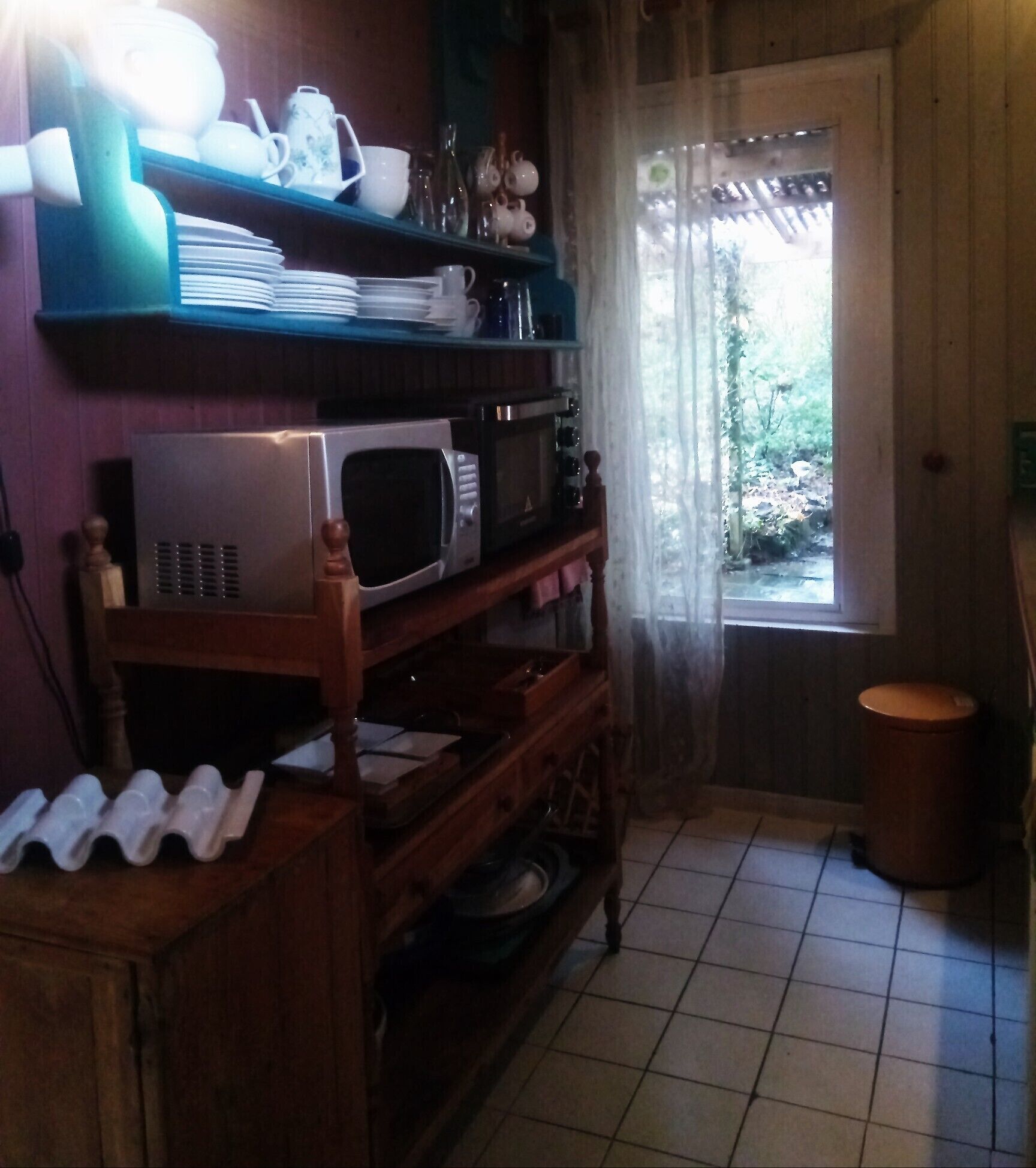 Une cuisine avec une étagère en bois où sont rangées différentes assiettes et un four à micro-ondes. Une fenêtre offre une vue sur la verdure à l’extérieur.