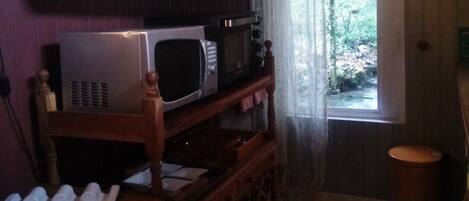 Une cuisine avec une étagère en bois où sont rangées différentes assiettes et un four à micro-ondes. Une fenêtre offre une vue sur la verdure à l’extérieur.