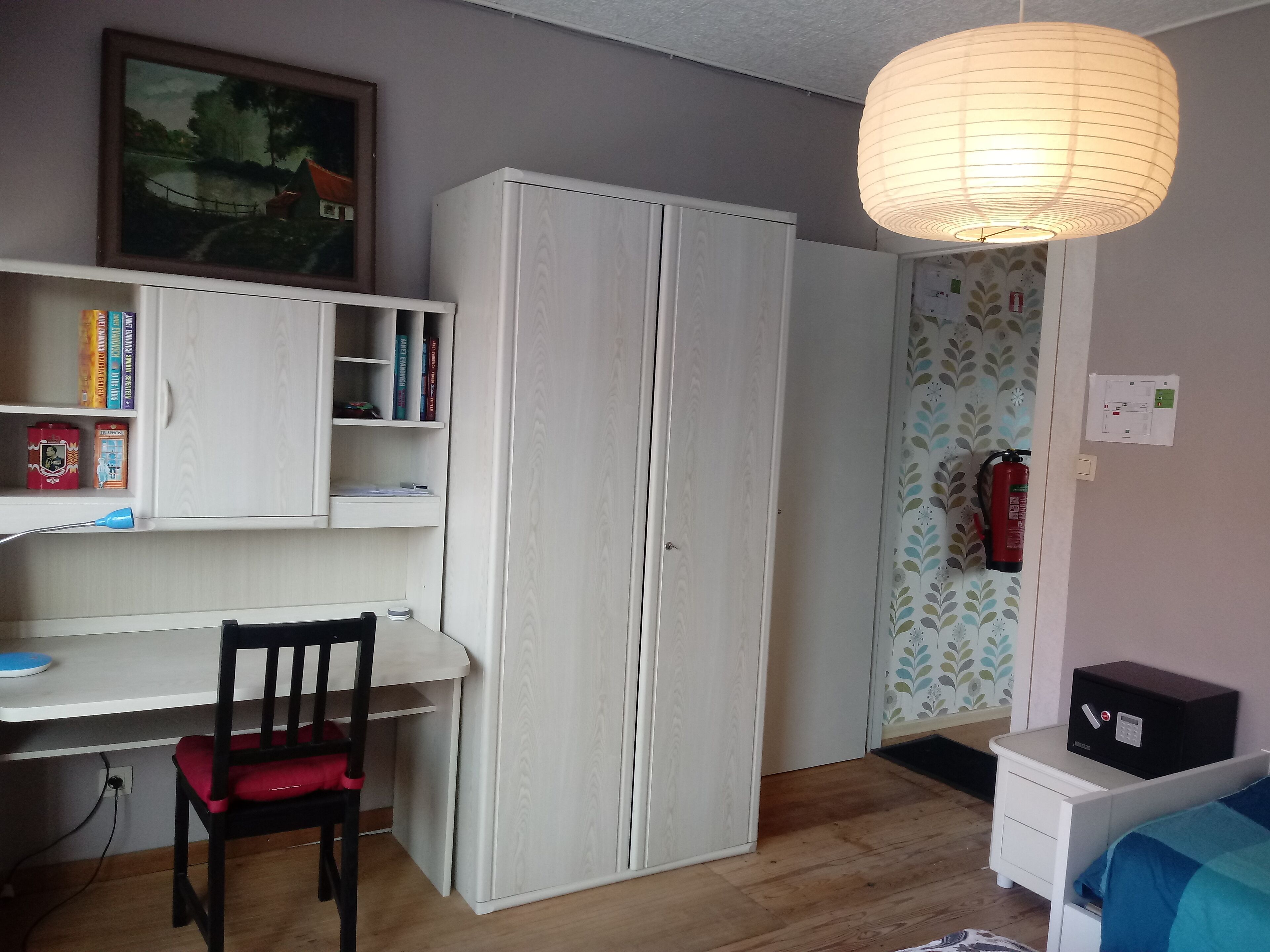 A bedroom with a bed, a desk with a chair, a wardrobe, a bookshelf, and a hanging light fixture.