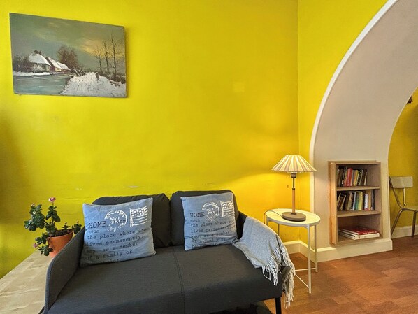 A room with a yellow wall, a sofa with two pillows, a small table with a lamp, a bookshelf, and a painting of a snowy landscape. - A vacation house for a family/ group of 4 in Ghent (Gent)