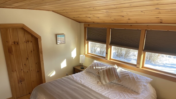 Een slaapkamer met een bed, houten plafond, een raam met zonwering, een nachtkastje en een lijst met een afbeelding aan de muur.