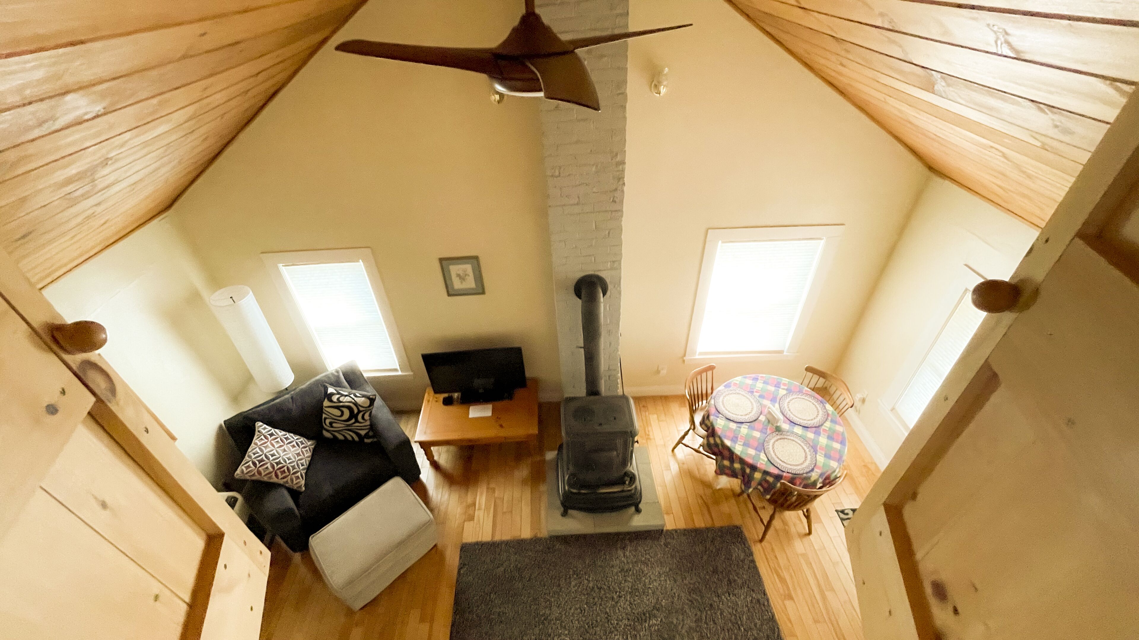 Un acogedor espacio tipo loft con techo inclinado, chimenea, una mesa de comedor con sillas, un sofá con cojines y un televisor sobre una mesa de madera.