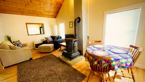 Een gezellige woonkamer met een open haard, een eettafel met een kleurrijk tafelkleed, en een comfortabele zithoek.