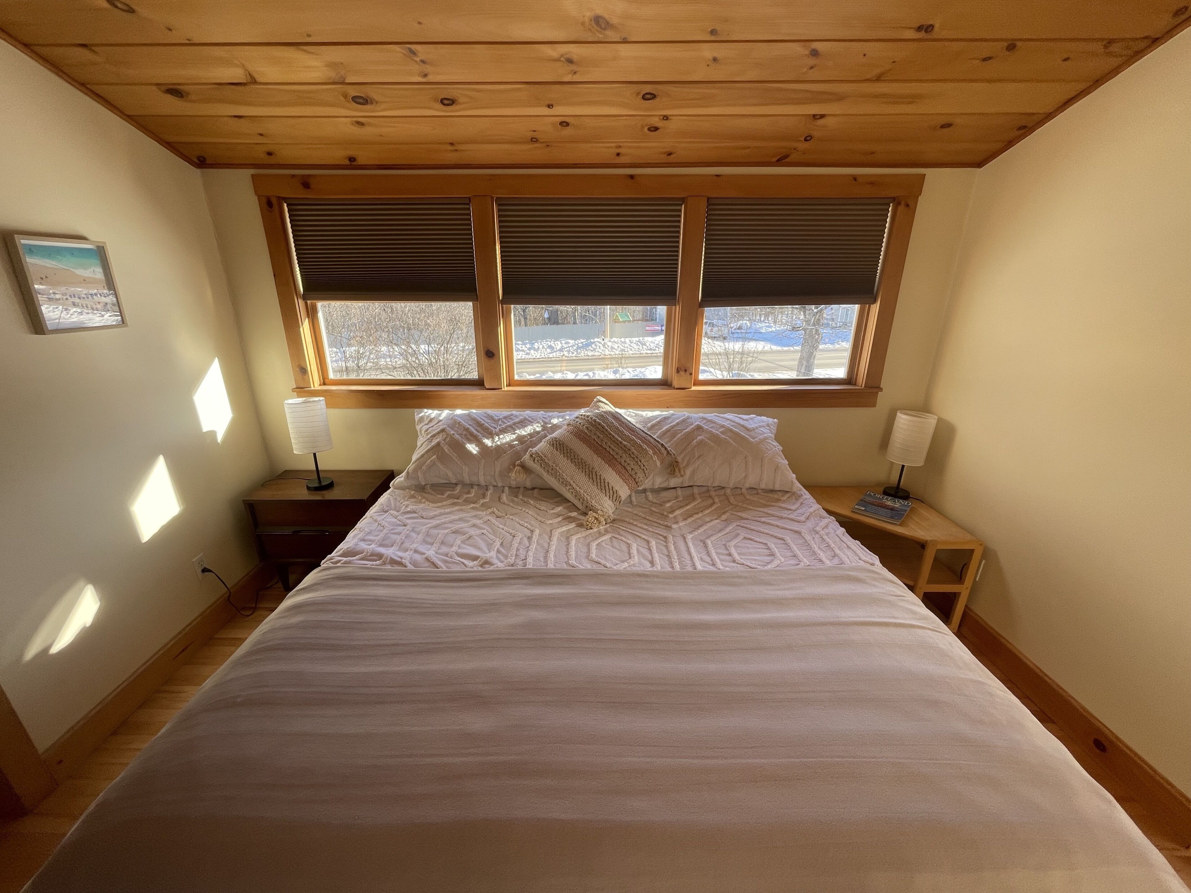 Un dormitorio con techo de madera, cama con ropa de cama blanca, dos mesitas de noche con lámparas y una ventana con persianas.