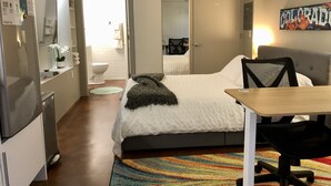 Une chambre d’hôtel comprenant un lit, un bureau, une chaise, un réfrigérateur, un lavabo, un miroir et une salle de bain visible à travers une porte ouverte.