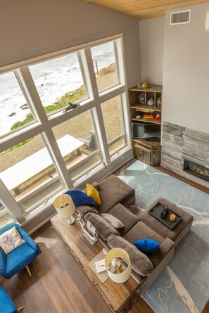 A living room with a large window overlooking the beach, a fireplace, and a cozy seating area. - Lookout House - Romantic Luxury and Ocean Views (Gold Beach)