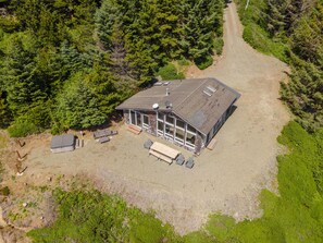 An aerial view of a cabin with a large deck, surrounded by dense forest. - Lookout House - Romantic Luxury and Ocean Views (Gold Beach)