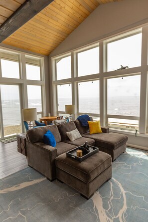 A spacious living room with a large sectional sofa, a coffee table, and a view of the ocean through large windows. - Lookout House - Romantic Luxury and Ocean Views (Gold Beach)