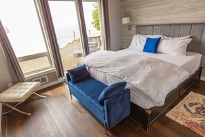 A hotel room with a bed, a blue chair, a window with a view of the sea, and a wooden floor.