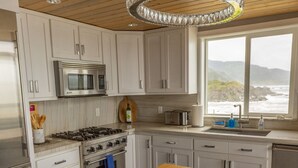 A modern kitchen with white cabinets, stainless steel appliances, a wooden island, and a view of the ocean through a large window. - Lookout House - Romantic Luxury and Ocean Views (Gold Beach)