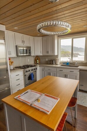 A modern kitchen with white cabinets, stainless steel appliances, a wooden island, and a view of the ocean through a large window. - Lookout House - Romantic Luxury and Ocean Views (Gold Beach)