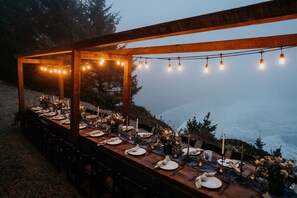 An outdoor dining setup with a wooden table, chairs, string lights, and floral arrangements.