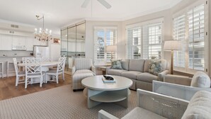 A spacious living area with a dining table, chairs, a sofa, and a coffee table. There is a ceiling fan, large windows with shutters, and a kitchen in the background. - 81 Ocean Place - Lovely Oceanfront 3rd Floor With Views Of The Atlantic Ocean (Fernandina Beach)
