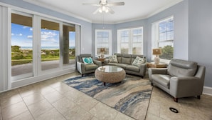 A spacious living room with a large sliding glass door leading to a balcony, a ceiling fan, and a round coffee table. - 5 Ocean Place -1st Floor Oceanfront 3 BR Condo Next Door to The Ritz Carlton. (Fernandina Beach)