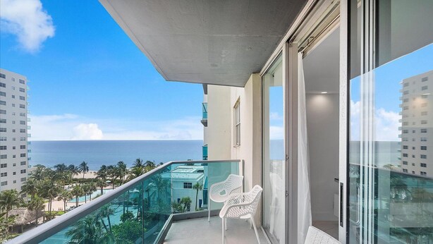 Un balcon doté d’une balustrade en verre, offrant une vue sur une piscine et l’océan, avec des chaises blanches et un ciel dégagé.