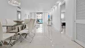 Un intérieur moderne avec une table à manger en verre, des chaises blanches, un lustre et une vue sur un immeuble à travers une porte vitrée.