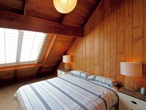 A bedroom with a sloped ceiling, wooden walls, a bed with striped bedding, two bedside tables with lamps, and a window with blinds. - Ti Kouka Chalet - Ohakune Holiday Home (Ohakune)
