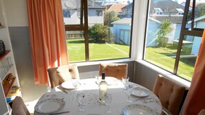 Un coin repas avec une table dressée pour quatre personnes, une bouteille d’eau et une vue sur une pelouse verdoyante et des maisons, le tout visible à travers de grandes fenêtres.