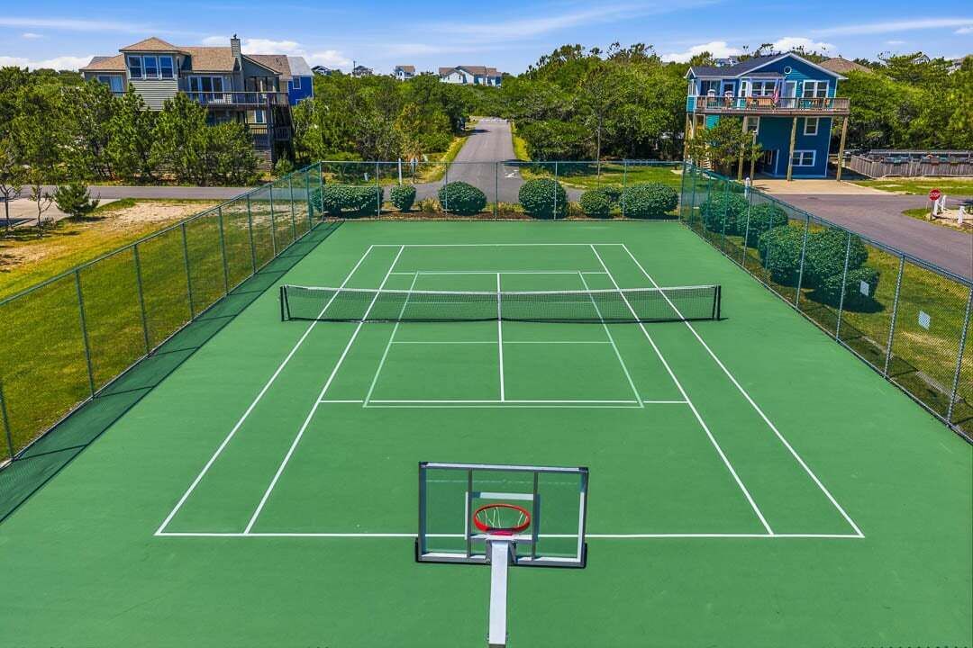En tennisbane med en basketballkurv, omkranset af et hegn, og huse i baggrunden.