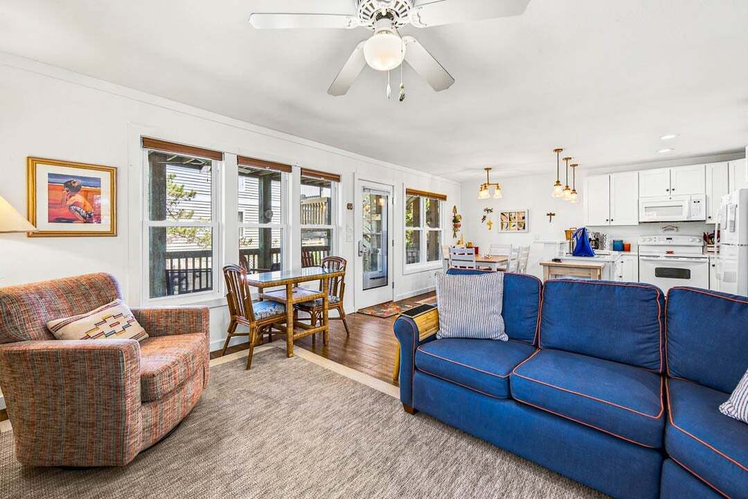 A living room with a blue sofa, a dining table with chairs, a ceiling fan, and a kitchen area with cabinets and appliances.