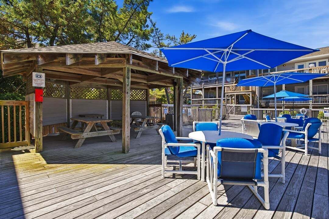 Una terraza de madera con mobiliario exterior y sombrillas, un espacio cubierto y un edificio al fondo.