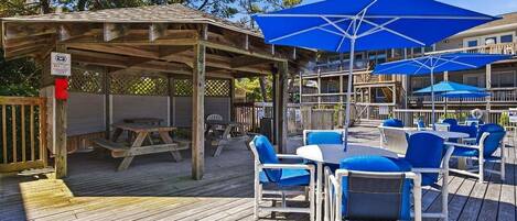 Una terraza de madera con mobiliario exterior y sombrillas, un espacio cubierto y un edificio al fondo.