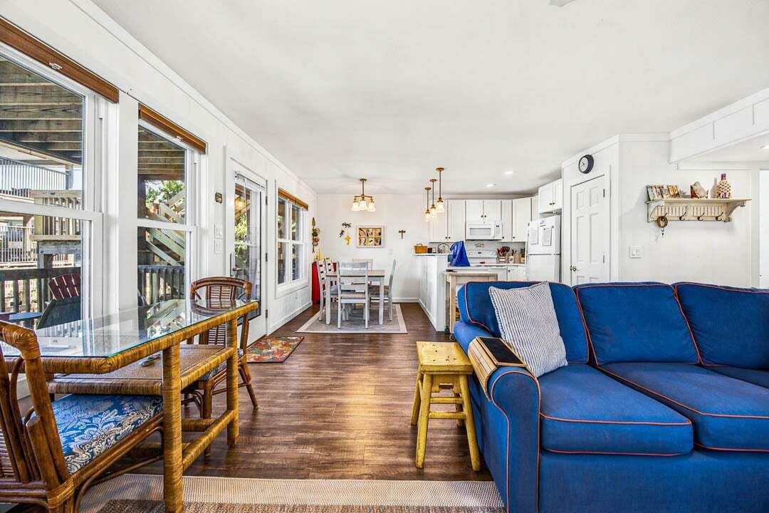 A spacious living area with a blue sofa, dining table, and chairs, a kitchen in the background, and large windows.