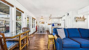 A spacious living area with a blue sofa, dining table, and chairs, a kitchen in the background, and large windows.