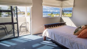 A bedroom with a bed, a desk, and two windows offering a view of the ocean and palm trees. - Sun, Sea, Sensational - Kaiteriteri Holiday Home (Kaiteriteri)