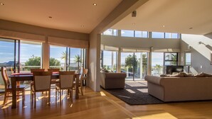 A spacious living room with a wooden floor, a sofa, a dining table with chairs, and large windows offering a view of the outdoors. - Sun, Sea, Sensational - Kaiteriteri Holiday Home (Kaiteriteri)