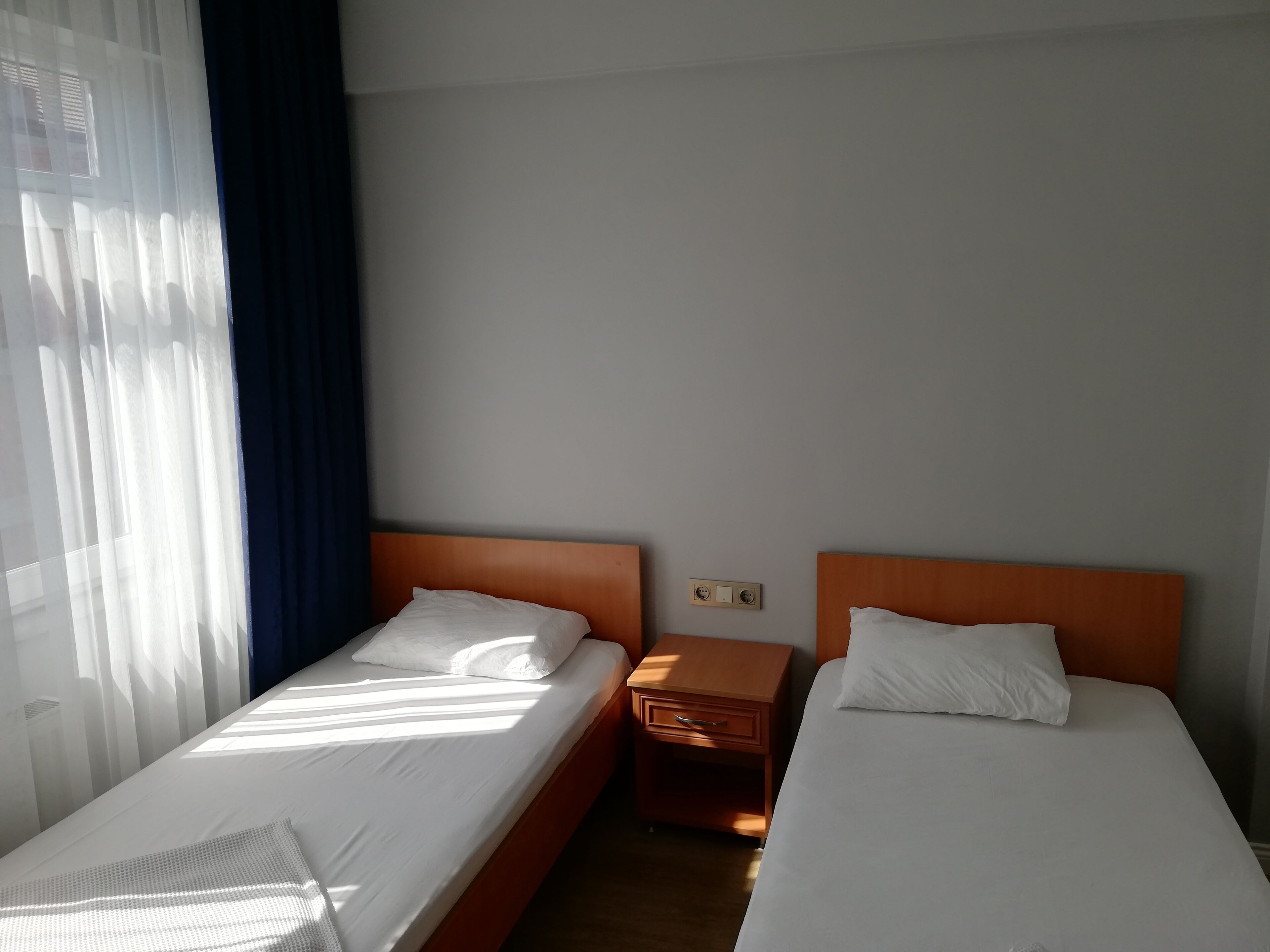 Una habitación de hotel con dos camas individuales, una mesita de noche de madera y una ventana con cortinas translúcidas.
