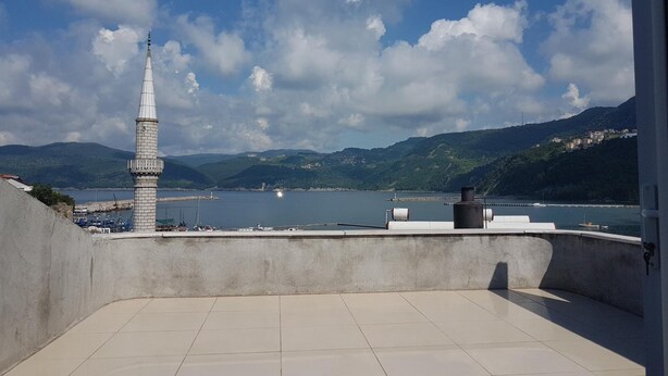 A terrace with a view of a body of water, a tower, and hills in the distance. - Karadeniz Pansiyon (Amasra)